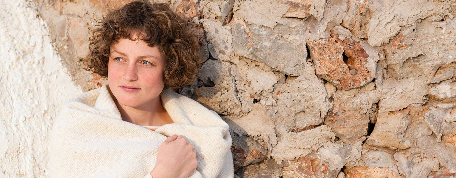 woman wrapped in a blanket, standing against a textured stone wall.