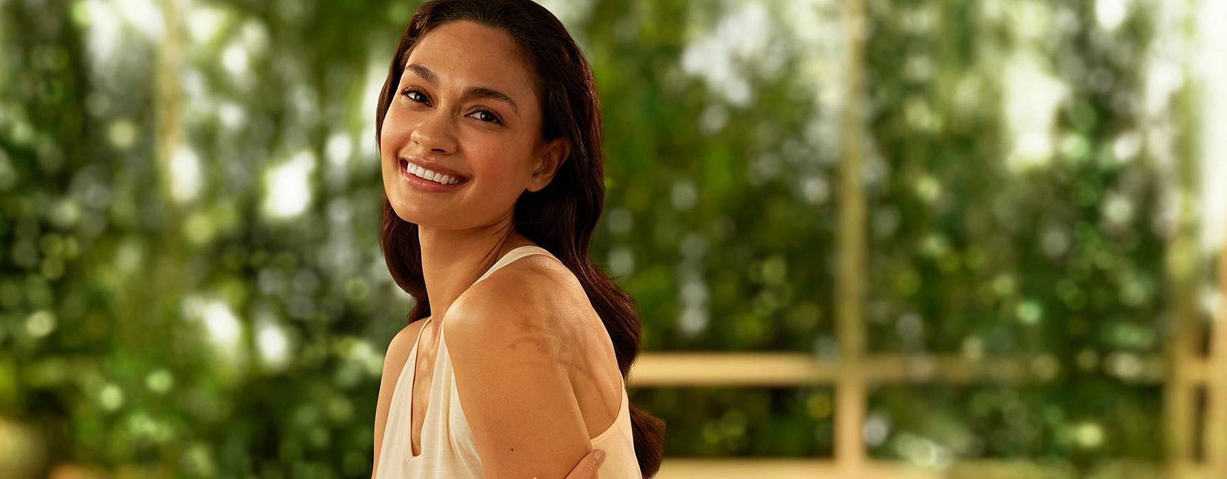 woman smiling, touching her arm in a softly lit, indoor setting with lush green plants in the background.