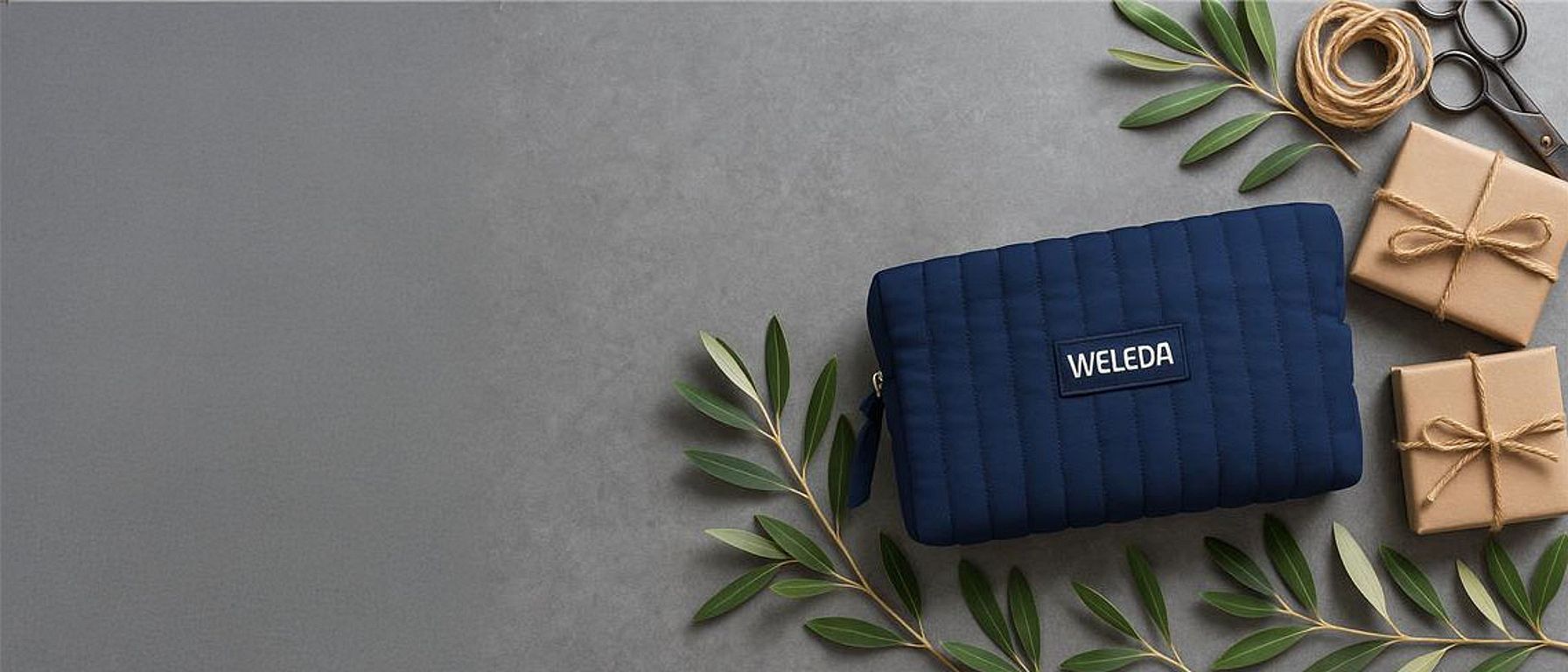 Blue Weleda Toiletry bag on a grey background surrounder by gifts and green leaves