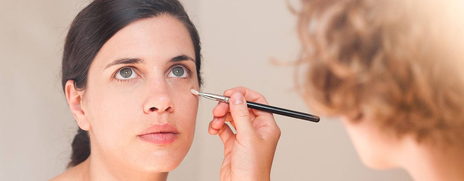 woman having makeup applied under her eye with a brush by another person.