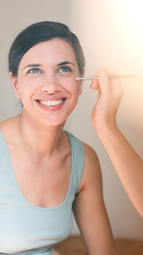 laughing woman having makeup applied under her eye with a brush by another person.