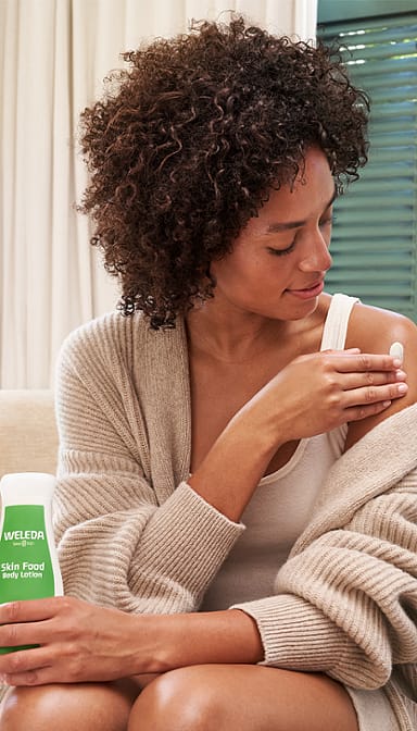 Dark skin woman holding and applying Skin Food Body Lotion to her shoulder.