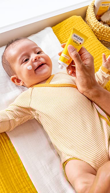Smiling baby wearng yellow, lying down and being applied Calendula Face Cream on his face.