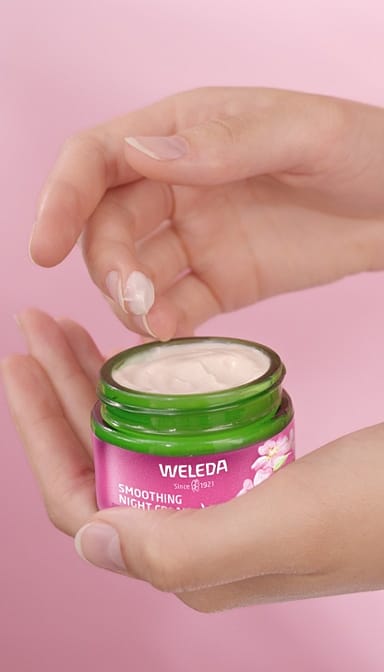 Woman's hand holding an open jar of Smoothing Night Cream Wild Rose & White Tea and dipping a finger into the jar.