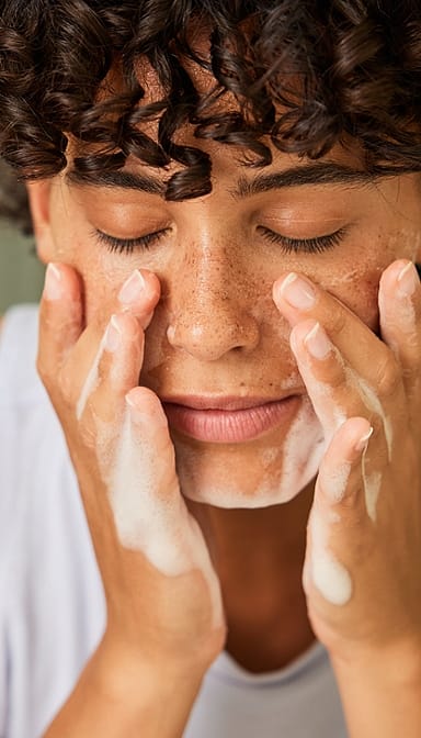 Dark skin woman with short curly hair with her eyes closed washing her face with a face cleasing foam.