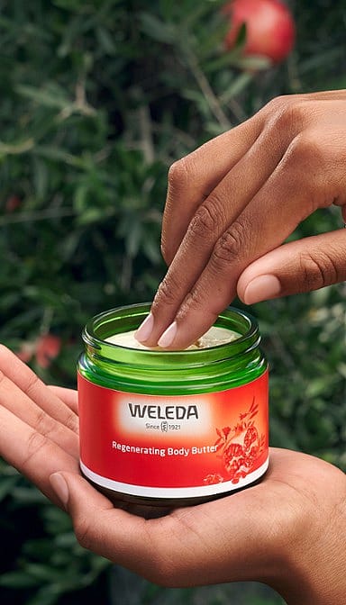 Woman holding open jar of Regenerating Body Butter Pomegranate and dipping fingers into yhe