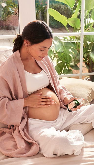 Dark hair woman sitting down and applying Stretch Mark Massage Oil to her pregnant belly.