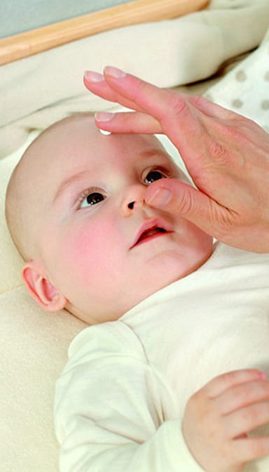 Baby lying down and hand applying cream to his face.