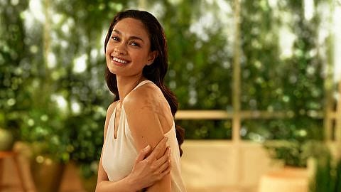 woman smiling, touching her arm in a softly lit, indoor setting with lush green plants in the background.