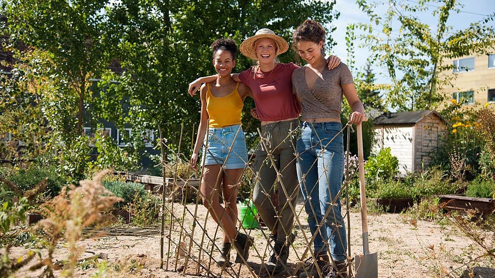 three women standing together outdoors in a garden, smiling and embracing each other proudly.