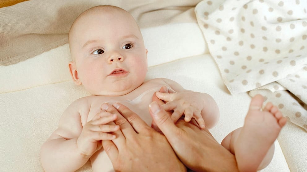 Baby lying down on whte linen being covered in body lotion.