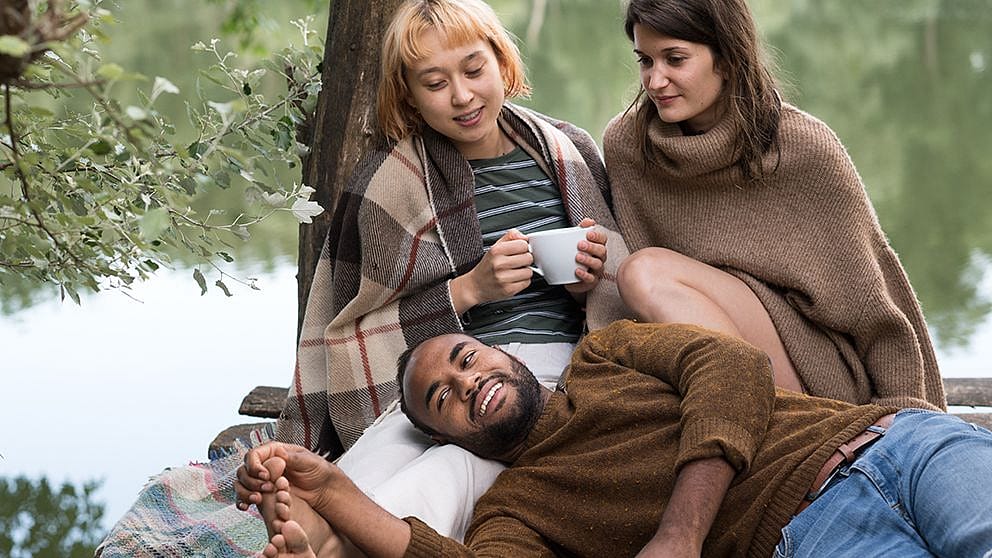 Two women and a man enjoying a cup of tea under a tree.