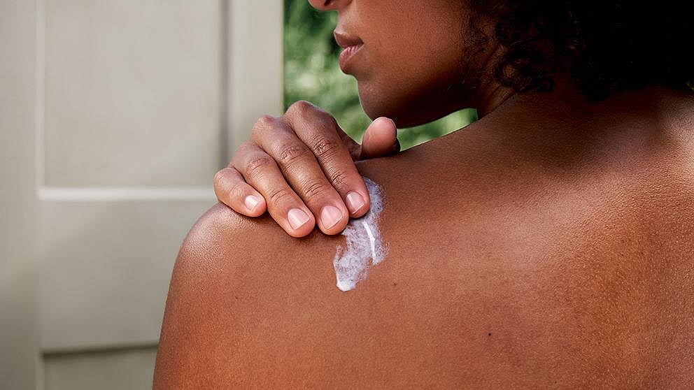 Dark skin woman applying Weleda Regenerating Body Butter onto her back.